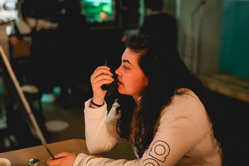 A young woman with long, curly dark hair is sitting at a table in a dimly lit indoor setting, speaking into a walkie-talkie. She is wearing a white long-sleeve shirt with a circular design on the sleeve. Her facial expression is focused and serious.