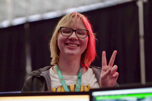 A young woman with short blonde hair and glasses smiles brightly while flashing a peace sign. She is wearing a lanyard with a badge, a white t-shirt with a graphic design, and a dark jacket.