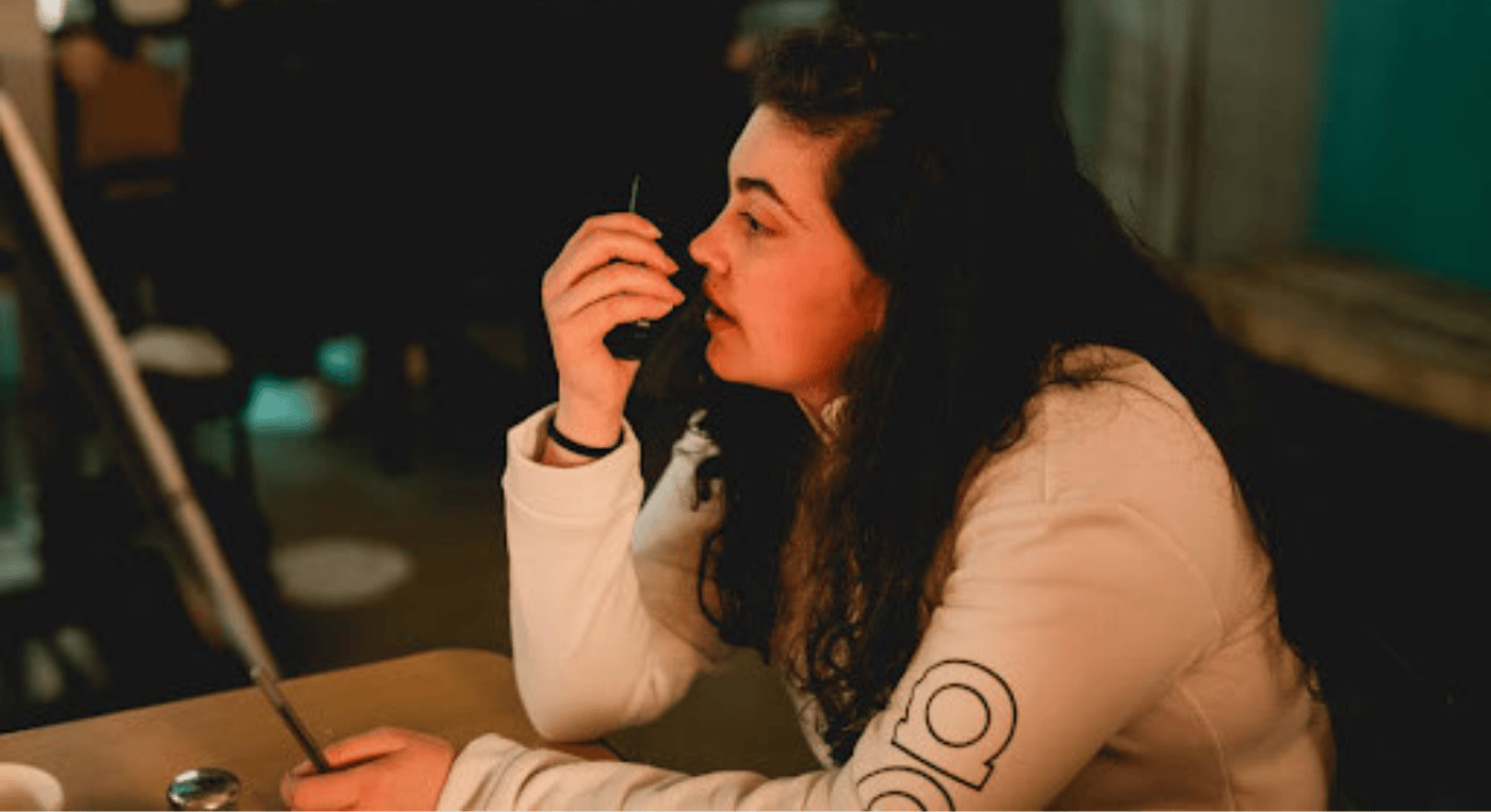 A young woman with long, curly dark hair is sitting at a table in a dimly lit indoor setting, speaking into a walkie-talkie. She is wearing a white long-sleeve shirt with a circular design on the sleeve. Her facial expression is focused and serious. The background is blurred, showing other people and a warmly lit environment, suggesting a workplace or event setting.