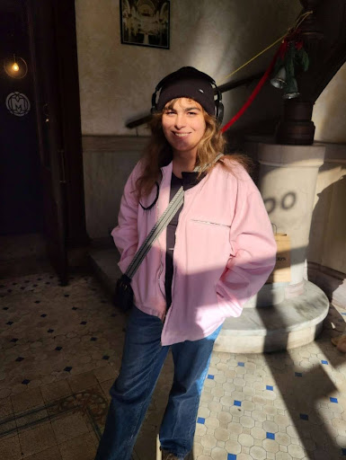 A young woman with long, wavy light brown hair stands in a sunlit indoor space, smiling slightly at the camera. She is wearing a black beanie, large over-ear headphones, a pink zip-up jacket, blue jeans, and a crossbody bag.