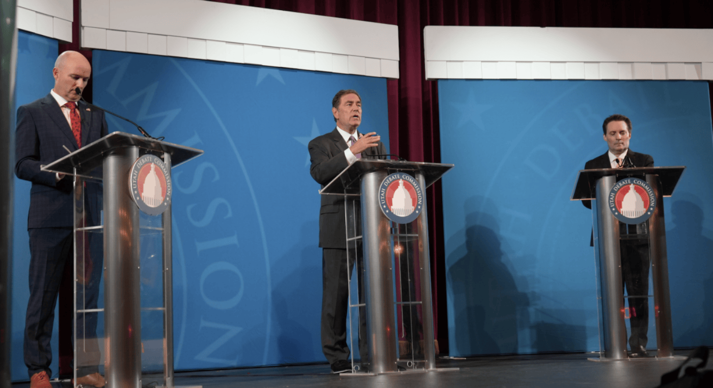 Three men standing behind clear podiums on a blue stage 
